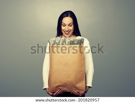 excited smiley businesswoman holding paper bag and looking at money. photo in studio over grey background