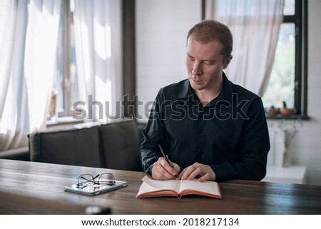 A young man writes in a notebook at the table. Journalist, writer makes notes, works on the text at the workplace in a bright room in the house