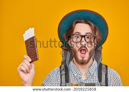 Tourism and travel, adventure. Funny young man with backpack and tourist hat surprises holding his passport and tickets looking at  camera. Studio portrait on a yellow background.