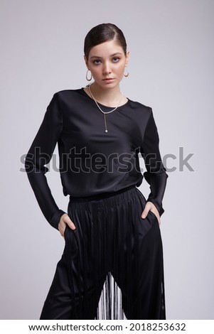 High fashion photo of a beautiful elegant young woman in a pretty black blouse, fringe trousers, accessories over gray background. Studio Shot. Gathered dark hair