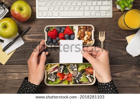 First person top view photo of hands holding white sticky note over two lunchboxes with healthy food glass of juice plant stationery keyboard mouse isolated dark wooden desk background with copyspace