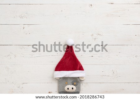 Children's knitted hat with a picture of a bull and wearing a Christmas hat of santa claus on the background of a white wooden table.