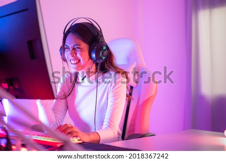 Happy young pretty Asian transgender professional gamer wearing a gaming headset raising fist up after winning the game while having fun playing online video games on a personal computer in bedroom.