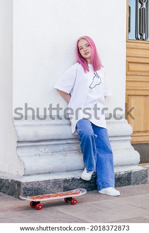 A pink-haired hipster teenage girl in a white T-shirt and jeans standing with a skateboard on a city street on a summer day.Generation Z style,active lifestyle.Copy space.