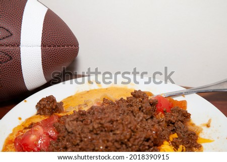 Spicy ground beef on a white plae with diced tomatoes and shredded cheese next to a football