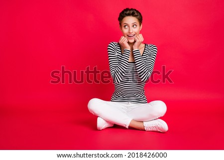 Photo of sweet impressed woman dressed striped outfit legs crossed arms cheeks looking empty space isolated red color background