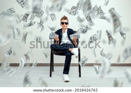 A rich boy with glasses sits on a chair and holds a bundle of dollars in his hands. Knowing how to make money. Money rain, Financial education, young millionaire, investments, loans, winnings