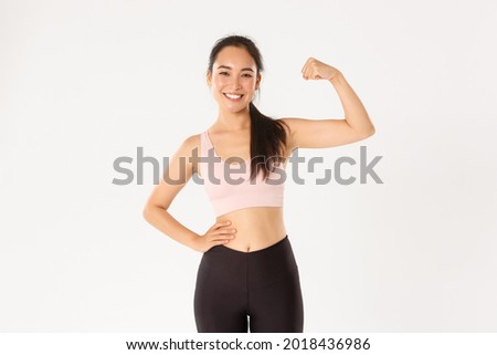 Sport, wellbeing and active lifestyle concept. Portrait of smiling slim and strong asian fitness girl, personal workout trainer showing muscles, flexing biceps and look proud, white background Royalty-Free Stock Photo #2018436986