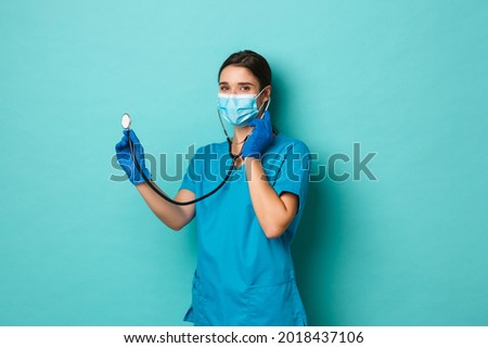 Covid-19, pandemic and medicine concept. Beautiful female doctor in medical mask, gloves and scrubs, having patient check up with stethoscope and smiling at camera, blue background
