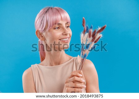 Joyful woman with pink hair smiling and holding dried flowers of neutral colors in her hand, posing over blue studio background. Reed stem, artificial nordic style dried flower composition for home