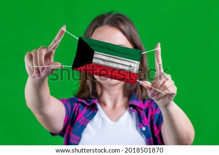 Woman in a plaid shirt holds a medical mask with of the Kuwait flag on a green background