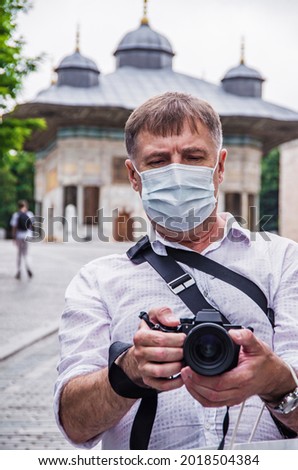 Portrait of a photographer, a man with a medical mask, holding a professional digital DSLR camera, enjoying his work outdoors.