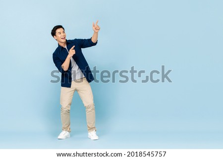 Full length portrait of smiling young handsome Asian man pointing fingers to empty space aside in isolated studio light blue background Royalty-Free Stock Photo #2018545757