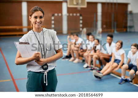 Young happy female coach looking at camera while having PE class with elementary students at school gym.  Royalty-Free Stock Photo #2018564699