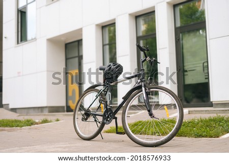 Modern bicycle standin on footplate in city street on blurred background of front door of apartment building, office building. Protective helmet lies on seat of bike, outdoors, no people, nobody. Royalty-Free Stock Photo #2018576933