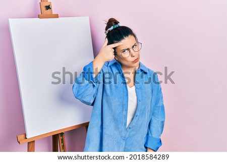 Young hispanic woman standing by painter easel stand pointing unhappy to pimple on forehead, ugly infection of blackhead. acne and skin problem 