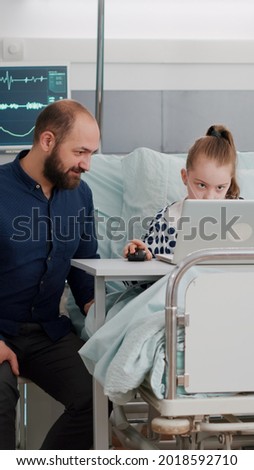 Father sitting beside sick daughter while playing cartoon online video games using laptop computer after suffering recovery surgery. Child sitting in bed during therapy consultation in hospital ward