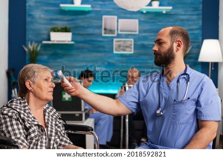 Assistant man helper checking temperature using medical infrared thermometer discussing with senior woman during therapy. Social services nursing elderly retired female. Healthcare assistance