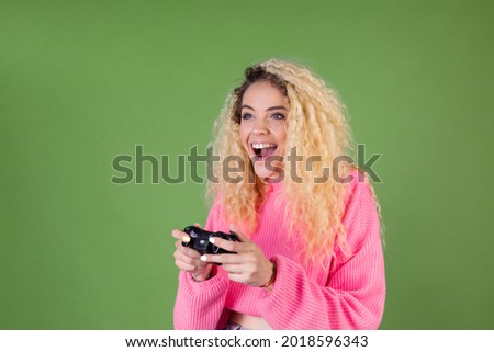 Young blonde woman with long curly hair in pink sweater on green background with joystick playing games