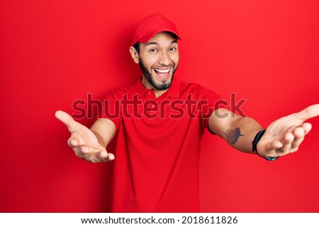 Hispanic man with beard wearing delivery uniform and cap smiling cheerful offering hands giving assistance and acceptance. 