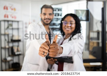 Blurred view of two cheerful multiracial doctors in white lab coats, showing thumbs up while standing together at medical center. Concept of people, medicine and teamwork. Focus on hands