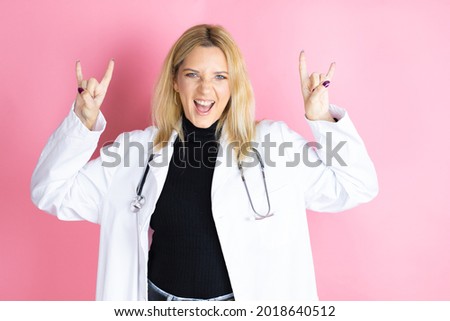 Young blonde doctor woman wearing stethoscope standing over isolated pink background shouting with crazy expression doing rock symbol with hands up