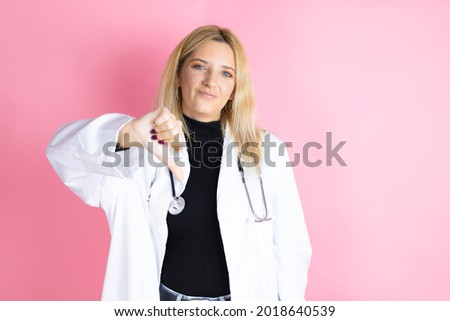 Young blonde doctor woman wearing stethoscope standing over isolated pink background with angry face, negative sign showing dislike with thumb down