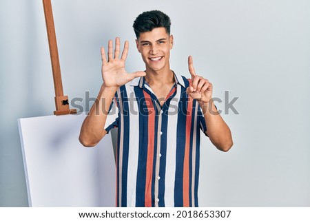 Young hispanic man standing close to empty canvas showing and pointing up with fingers number six while smiling confident and happy. 