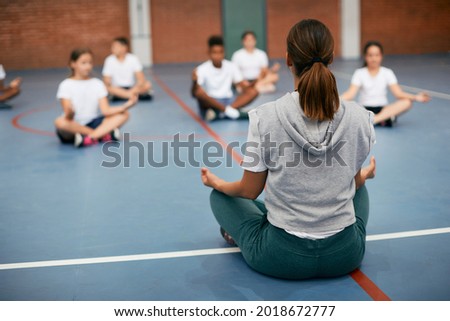 Back view of PE teacher leading Yoga class and meditating with elementary students at school gym. Royalty-Free Stock Photo #2018672777