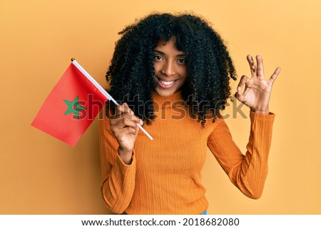 African american woman with afro hair holding morocco flag doing ok sign with fingers, smiling friendly gesturing excellent symbol 