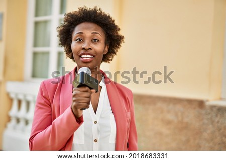Young african american woman journalist holding reporter microphone speaking and smiling to the camera for television news Royalty-Free Stock Photo #2018683331