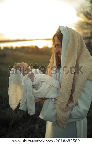 Saint Joseph holds baby Jesus in his arms and looks to him with love against meadow and river background.