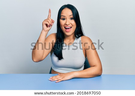 Beautiful hispanic woman with nose piercing sitting on the table pointing finger up with successful idea. exited and happy. number one. 