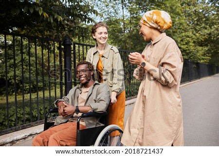 Young multi-ethnic friends talking during stroll: Caucasian woman pushing wheelchair with disabled man while talking to Arabian friend Royalty-Free Stock Photo #2018721437