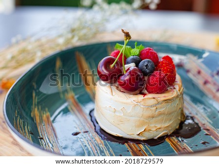 Summer dessert for serving in a restaurant. Meringue dessert with fresh cherries, raspberries, strawberries, blueberries and lemon balm, in a ceramic plate on a natural wood background.