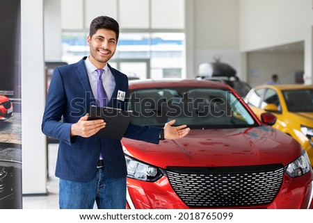 Handsome middle-eastern arab man sales manager showing brand new automobile in luxury auto showroom, holding chart, giving information about nice red automobile, copy space. Car retail concept Royalty-Free Stock Photo #2018765099