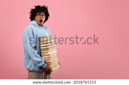 Nerdy black teenage student feeling shocked over pile of homework, holding big stack of books on pink background, banner with free space. College education, university exam concept