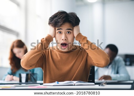 Portrait of shocked asian male student sitting at desk in classroom, grabbing his head looking at camera. Worried youth unprepared for test or exam, thinking about deadline or hard new theme Royalty-Free Stock Photo #2018780810
