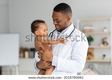 Portrait Of Handsome Young Black Pediatrician Holding Adorable Little Infant Boy In Hands, Cute Toddler Child Playing With Stethoscope On Doctor's Neck While Having Medical Check Up In Clinic Royalty-Free Stock Photo #2018780882