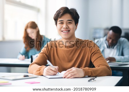 Modern Education Concept. Portrait of smiling asian male student sitting at desk in classroom at university, taking test or writing notes in his notebook, looking posing at camera, selective focus Royalty-Free Stock Photo #2018780909