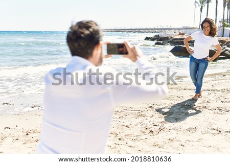 Middle age hispanic man make photo by the smartphone to woman at the beach.