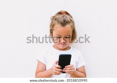 Little blond hair girl wearing white t-shirt, looking at mobile phone, with concentrated face