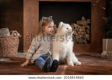 Beautiful little blonde baby girl, has happy fun face, pretty eyes, embrace samoyed husky dog puppy. Portrait holiday home. Winter family animal. Kids fashion style. 