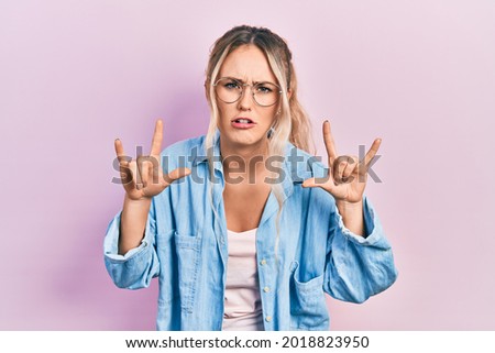 Beautiful young blonde woman wearing casual clothes and glasses shouting with crazy expression doing rock symbol with hands up. music star. heavy concept. 