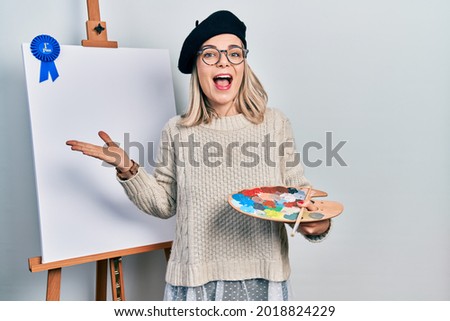 Beautiful caucasian woman drawing with palette on easel stand winner first place celebrating victory with happy smile and winner expression with raised hands 