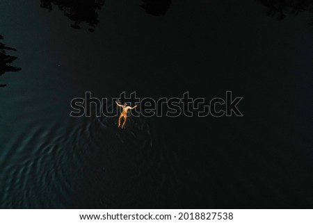 Aerial view above a woman swimming, in a forest pond, colorful, summer sunset, in Finland - top down, drone shot