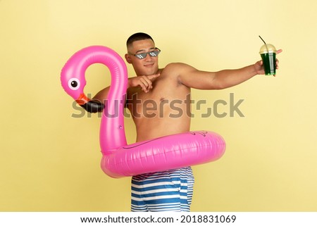 Drinking cool cocktail. Portrait of young handsome asian man in swimming trunks with pink inflatable flamingo circle isolated over yellow studio background. Concept of human emotions, leisure, youth.