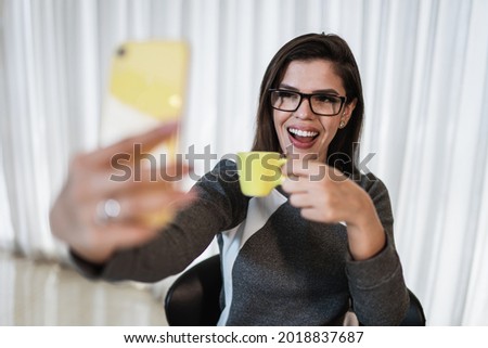 Portrait of a cheerful Latin girl dressed in grey clothes holding yellow coffee cup while taking a selfie at home