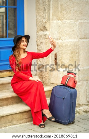 Tourist young caucasian woman in a red long dress and black hat with suitcase takes a selfie sitting on the stairs at the door outdoors