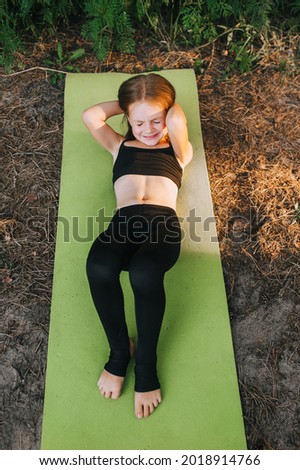A hardworking, preschool girl, child, a professional athlete, lies on her back, performs sports exercises, yoga and gymnastics, on a green rug, shaking her abdominal muscles. Photography, concept.